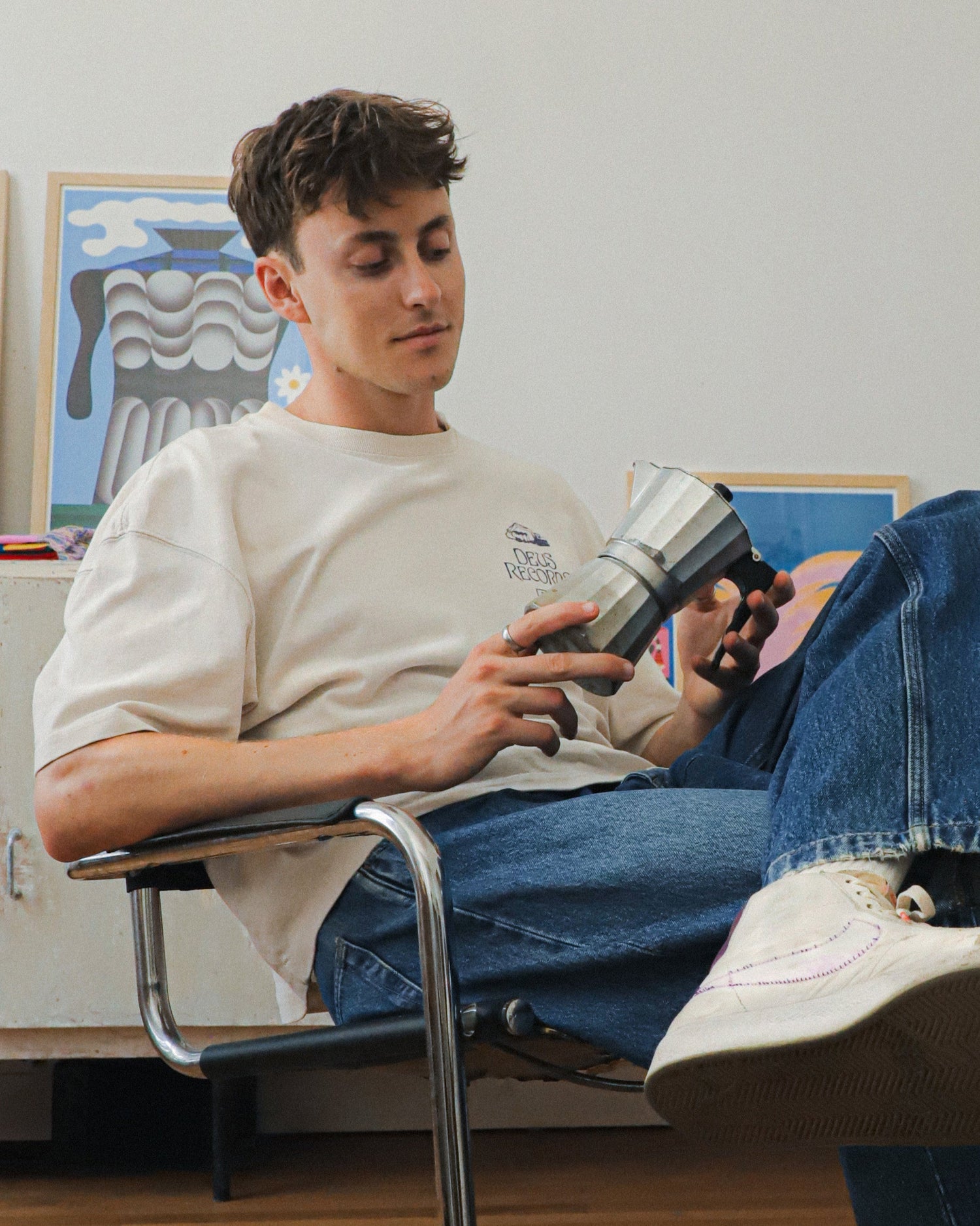 Person sitting on a chair holding a coffe pot in a room with artwork on the wall.